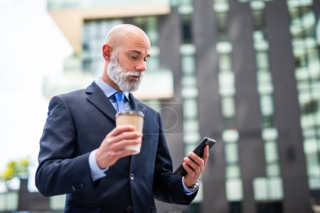 Foto de Un hombre de negocios calvo con barba blanca hablando por teléfono y bebiendo café - Imagen libre de derechos