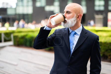 Téléchargez les photos : Homme d'affaires chauve avec une barbe blanche parlant boire du café tout en marchant dans une rue de la ville - en image libre de droit