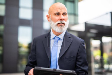 Téléchargez les photos : Homme d'affaires chauve à barbe blanche utilisant sa tablette dans une ville moderne - en image libre de droit