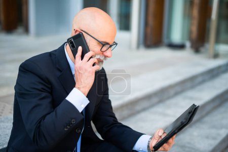 Foto de Hombre de negocios calvo con barba blanca usando su tableta mientras habla por teléfono en una ciudad moderna - Imagen libre de derechos