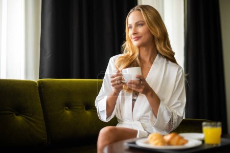 Téléchargez les photos : Jeune femme blonde boire une tasse de café assis sur un fauteuil à la maison. - en image libre de droit