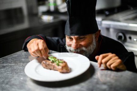 Photo for Cheerful white bearded chef garnishing a steak with rosemary - Royalty Free Image