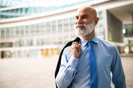 Téléchargez les photos : Mature chauve élégant portrait d'homme d'affaires avec une barbe blanche en plein air tenant sa veste - en image libre de droit