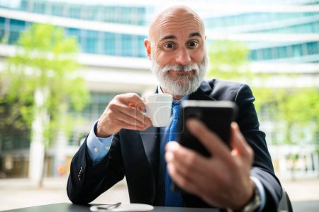 Photo for Senior stylish bald manager with white beard having a coffee outdoor while using his smartphone with a shocked expression - Royalty Free Image