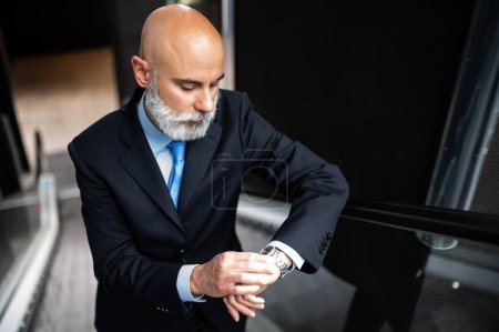 Photo for Mature bald stylish business man portrait with a white beard on mobile stairs holding his watch to check time - Royalty Free Image