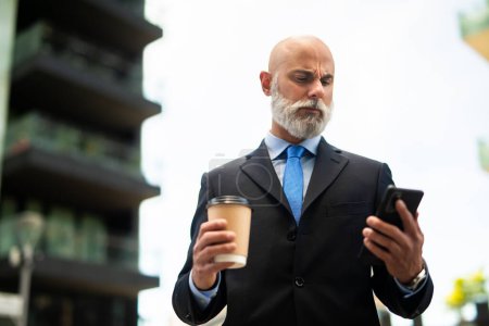 Foto de Elegante gerente calvo senior con barba blanca usando su teléfono inteligente al aire libre mientras bebe un café - Imagen libre de derechos