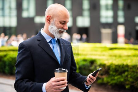 Photo for Stylish senior bald manager whit white beard using his smartphone outdoor while drinking a coffee - Royalty Free Image