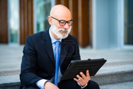 Foto de Administrador elegante usando su tableta sentada en un banco al aire libre en una ciudad moderna - Imagen libre de derechos
