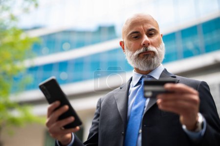 Photo for Handsome stylish bald manager using a smartphone outdoor to buy something with his credit card - Royalty Free Image