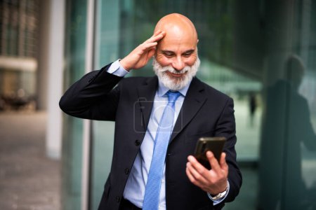 Téléchargez les photos : Happy senior chauve gestionnaire élégant avec barbe blanche dans une expression heureuse surprise shile lire des nouvelles sur son smartphone - en image libre de droit
