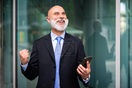 Photo for Successfull businessman in a victory pose outdoor while holding his smartphone - Royalty Free Image