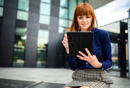 Foto de Sonriente pelirroja empresaria usando una tableta digital al aire libre sentada en un banco - Imagen libre de derechos
