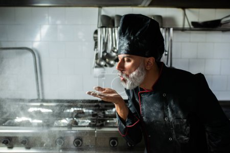 Téléchargez les photos : Un chef avec une barbe et un chapeau noir souffle de la farine dans sa bouche - en image libre de droit