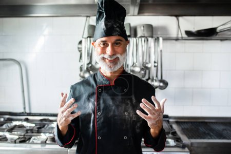 Foto de Un chef con barba y sombrero negro sonríe y extiende las manos - Imagen libre de derechos