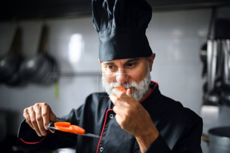 Experienced chef with gray beard in professional attire tasting food with focus and expertise