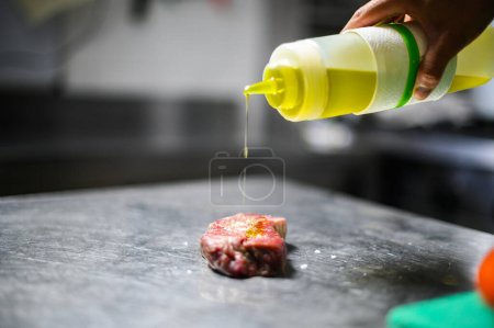 Chef's hand applying oil from a squeeze bottle to a raw steak on a steel countertop