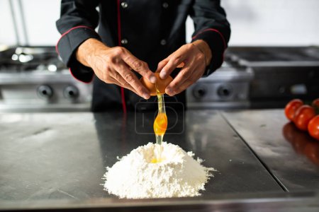 Téléchargez les photos : Chef prépare la pâte à pâtes fraîches en ajoutant un oeuf à un puits de farine sur un comptoir de cuisine - en image libre de droit