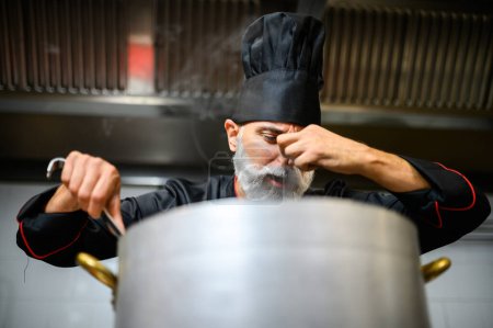 Foto de Chef masculino enfocado con barba degustando comida de una olla grande en una cocina industrial - Imagen libre de derechos