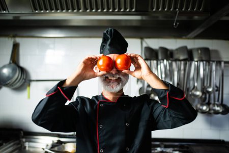 Photo for Cheerful chef holds tomatoes over his eyes, standing in a professional kitchen setting - Royalty Free Image