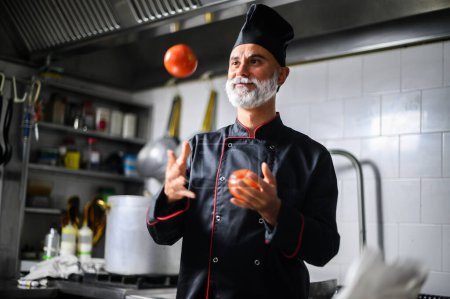 Photo for Cheerful male chef entertaining with a tomato juggling act in a professional kitchen environment - Royalty Free Image