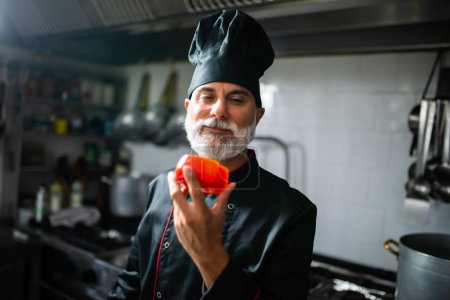 Foto de Chef profesional sonriente en uniforme mostrando pimiento fresco en una cocina comercial - Imagen libre de derechos