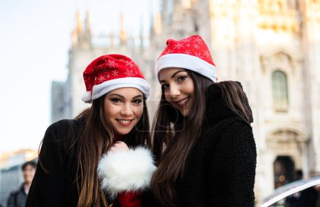 Photo for Two attractive smiling young women are shopping in Milan wearing Christmas hats - Royalty Free Image