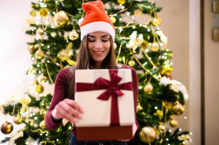 Photo for Young woman in a santa hat smiles while holding a festive gift in front of a decorated christmas tree - Royalty Free Image