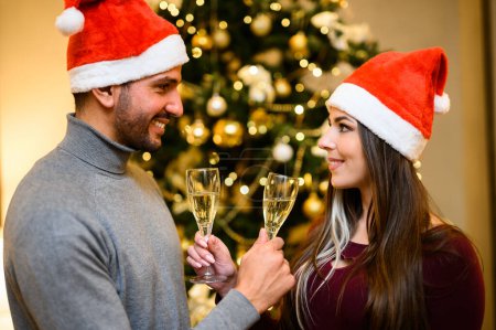 Foto de Alegre hombre y mujer en sombreros de santa tostada con champán por un árbol de Navidad - Imagen libre de derechos