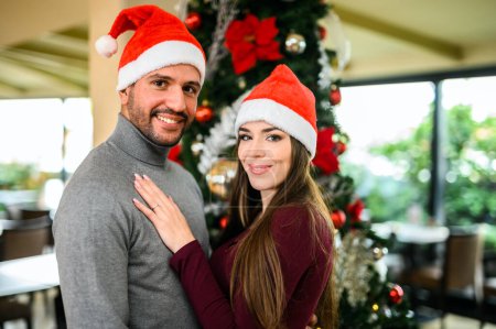 Photo for A diverse. Multi-ethnic couple embracing and celebrating christmas together with joyful smiles. Wearing santa hats. Surrounded by festive decorations and a cozy christmas tree - Royalty Free Image