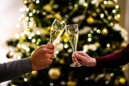 Photo for Close-up of hands clinking champagne glasses to celebrate, with a beautifully decorated christmas tree in the background - Royalty Free Image