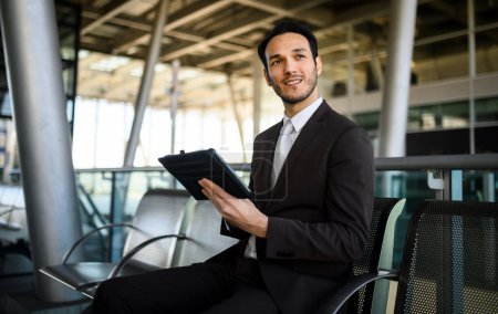 Foto de Joven hombre profesional en un traje trabajando en una tableta mientras espera en un aeropuerto - Imagen libre de derechos
