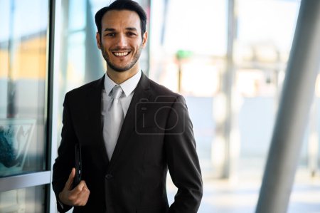 Foto de Hombre profesional en un traje con una sonrisa amistosa que sostiene a un planificador en un entorno corporativo moderno - Imagen libre de derechos