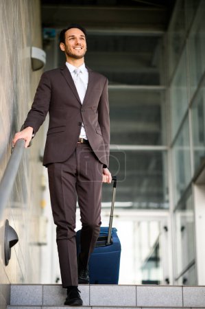 Téléchargez les photos : Jeune homme en costume souriant tout en marchant en bas devant un bâtiment moderne - en image libre de droit