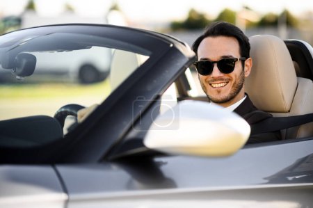 Téléchargez les photos : Heureux jeune homme adulte en lunettes de soleil conduisant une voiture convertible élégant par une journée ensoleillée - en image libre de droit
