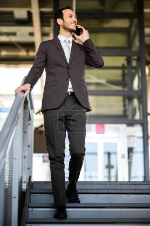 Foto de Alegre profesional masculino habla en su teléfono celular mientras está de pie en los escalones fuera de un edificio moderno - Imagen libre de derechos