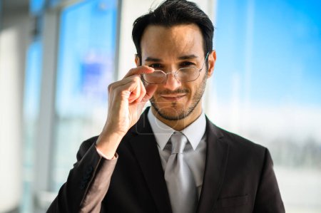 Foto de Hombre de negocios adulto joven serio ajustando gafas con estilo con confianza en un entorno de oficina corporativa moderna - Imagen libre de derechos