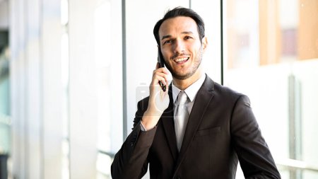 Photo for Confident young male professional in a suit talks on mobile phone indoors - Royalty Free Image