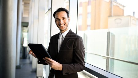 Téléchargez les photos : Homme professionnel souriant en costume tenant une tablette numérique dans un cadre urbain moderne - en image libre de droit