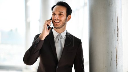 Foto de Confiado joven profesional masculino comprometido en una conversación en su teléfono móvil, de pie junto a una ventana - Imagen libre de derechos