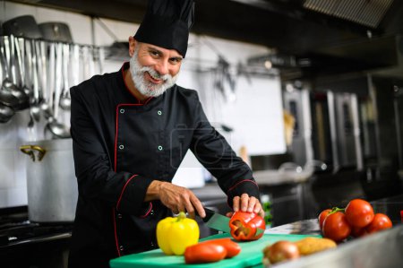 Foto de Chef macho sonriente en uniforme picando verduras frescas en una tabla de cortar - Imagen libre de derechos