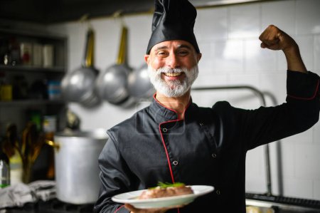 Foto de Chef sonriente en músculos uniformes de flexión mientras sostiene un plato de comida en una cocina profesional - Imagen libre de derechos