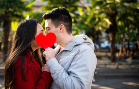 Photo for Young couple kissing and covering themselves with a heart shaped cardboard - Royalty Free Image