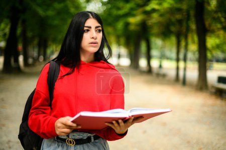 Photo for Focused young woman in a red hoodie reads a book while walking in a lush park - Royalty Free Image