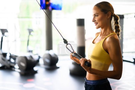Foto de Mujer joven enfocada en ropa deportiva utiliza una máquina de cable para el entrenamiento de fuerza en un gimnasio bien equipado - Imagen libre de derechos