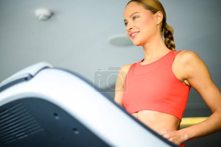 Foto de Mujer atlética joven en equipo de entrenamiento se centra en su carrera en una cinta de correr moderna en un entorno de gimnasio - Imagen libre de derechos