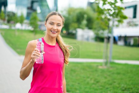 Photo for Smiling woman in sportswear holding a water bottle in an urban park setting - Royalty Free Image