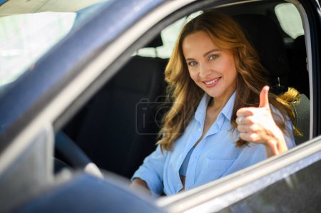 Téléchargez les photos : Femme souriante montrant son approbation avec un geste de pouce levé alors qu'elle était assise sur le siège du conducteur d'une voiture - en image libre de droit