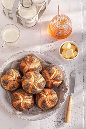 Tasty Kaiser buns with poppy seed served with milk. Breakfast with buns, butter and honey.