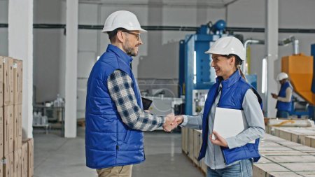 Photo for Two Caucasian workers wearing uniforms and helmets while standing in large storage or industrial facility. People actively talking together about distributing production. Using technology devices. - Royalty Free Image