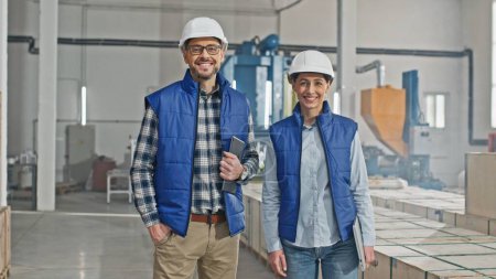 Photo for Caucasian engineers with safety helmets cheerfully smiling while looking at camera. Workers holding tablet device and laptop. Enjoying working at industrial facility. Around people located crates. - Royalty Free Image